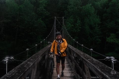A rider in a yellow jacket walking a suspension footbridge