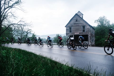 A pack of riders on a wet road passing an old clapboard building